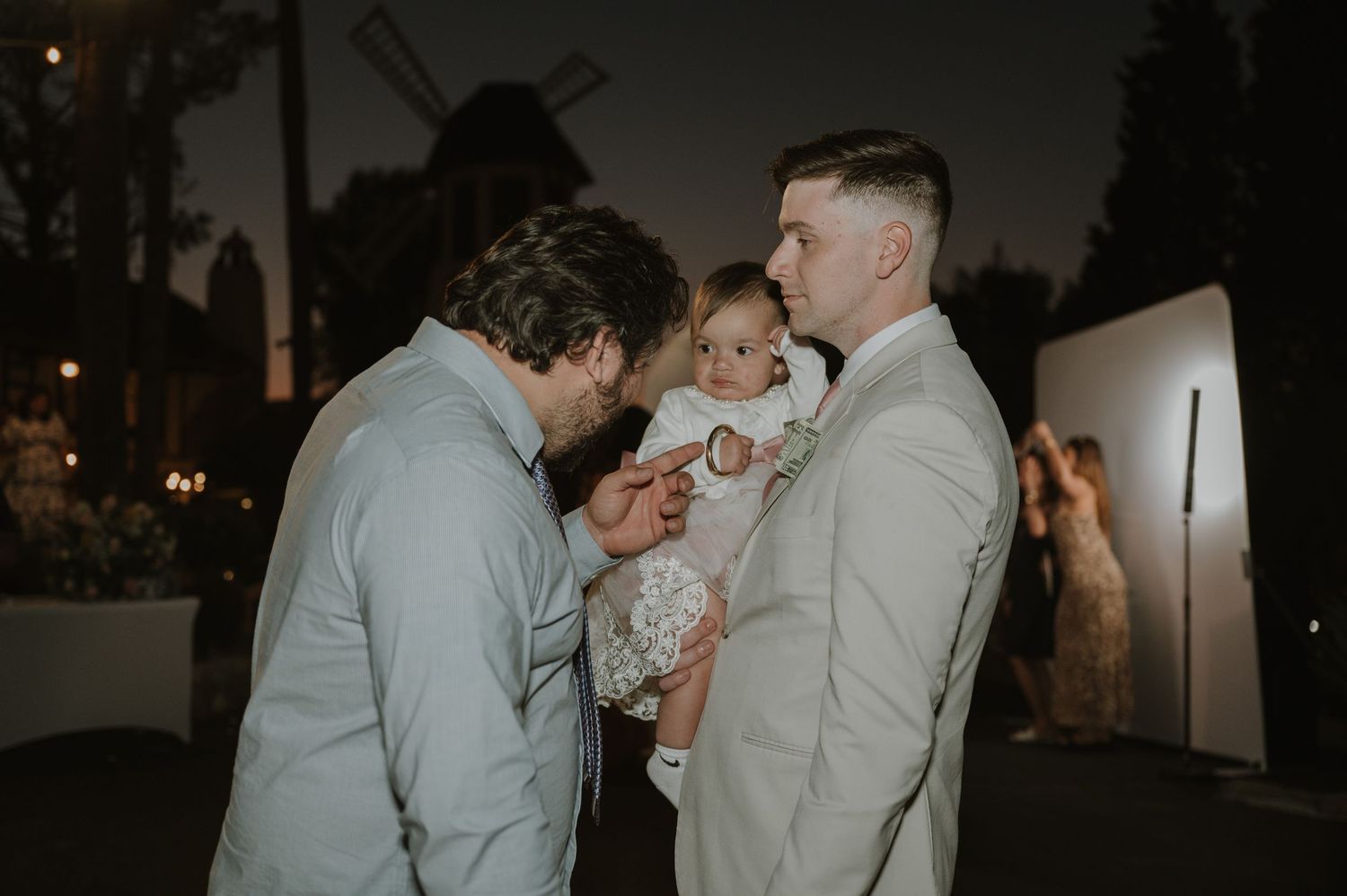 Two men admiring a baby in a white dress at an outdoor evening wedding reception.