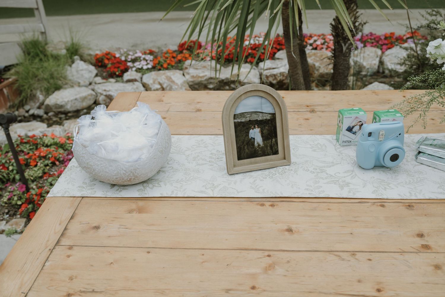 Rustic wooden table with a white vase, framed photo, and light blue Instax camera on a lace runner at an outdoor wedding.