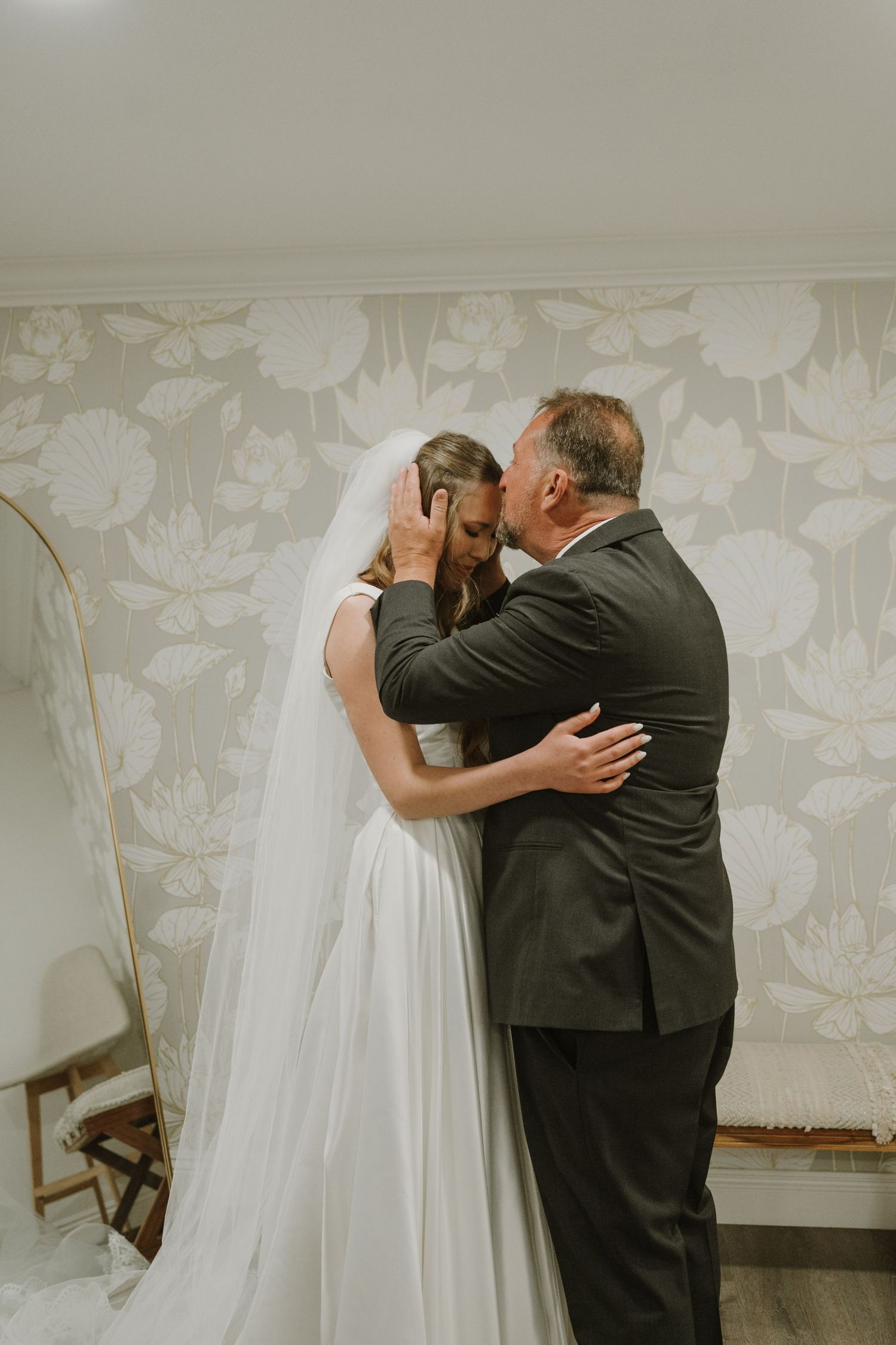 Emotional father kissing bride on forehead in room with floral wallpaper on wedding day.