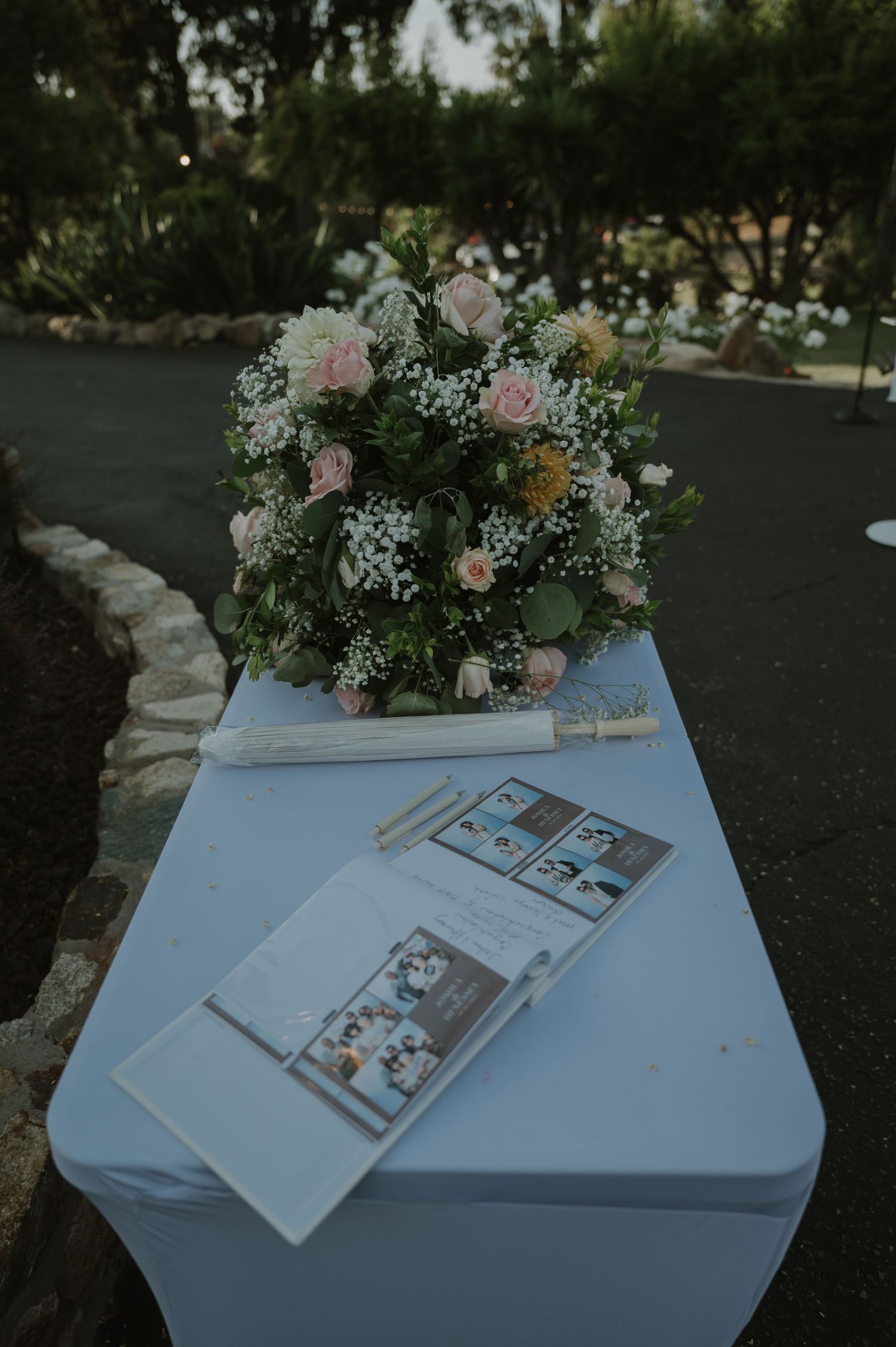Floral wedding bouquet with pink roses and baby's breath beside ceremony programs on a white draped table outdoors.
