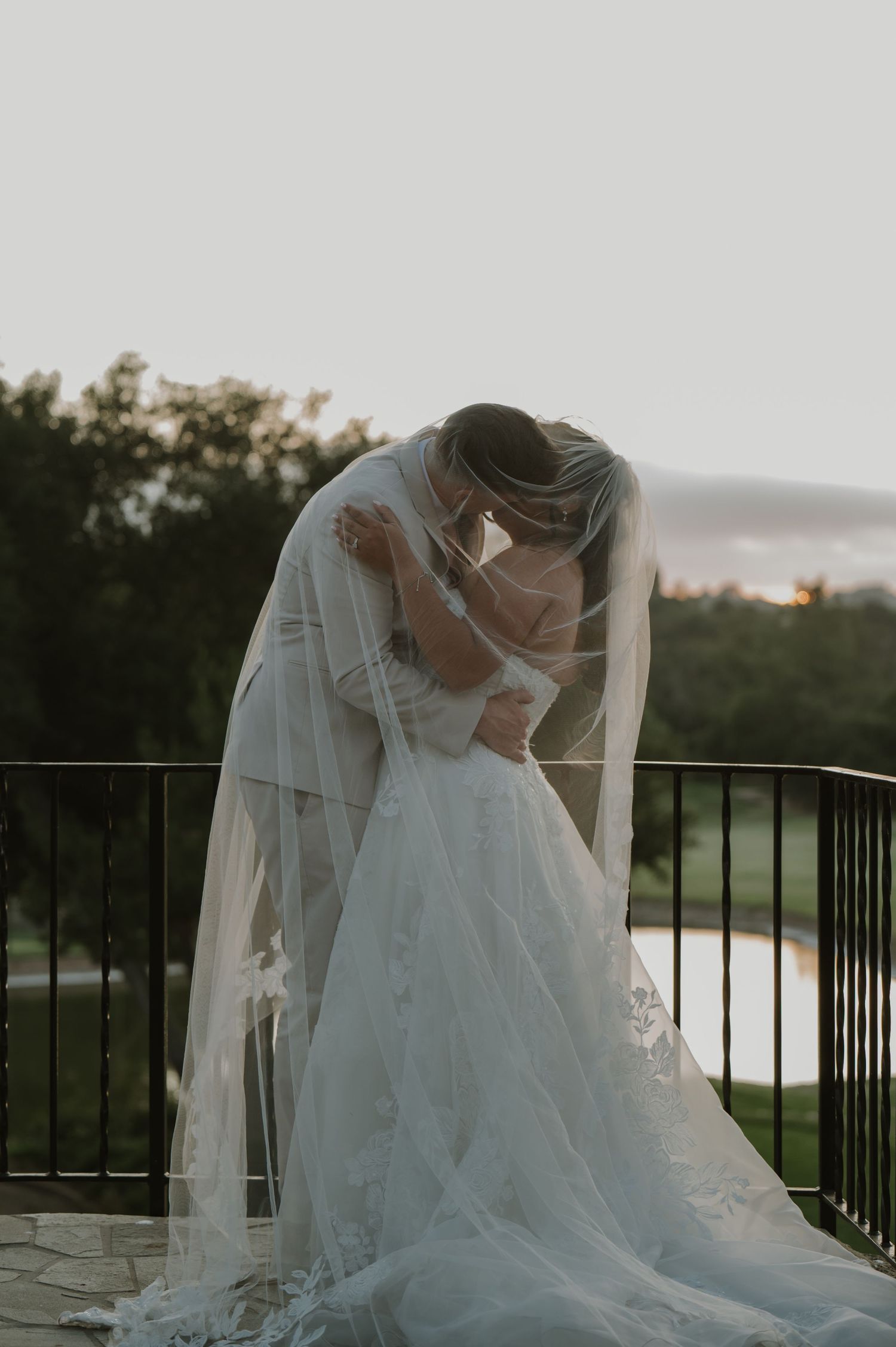 Bride and groom kiss under a long flowing veil on a balcony at sunset.