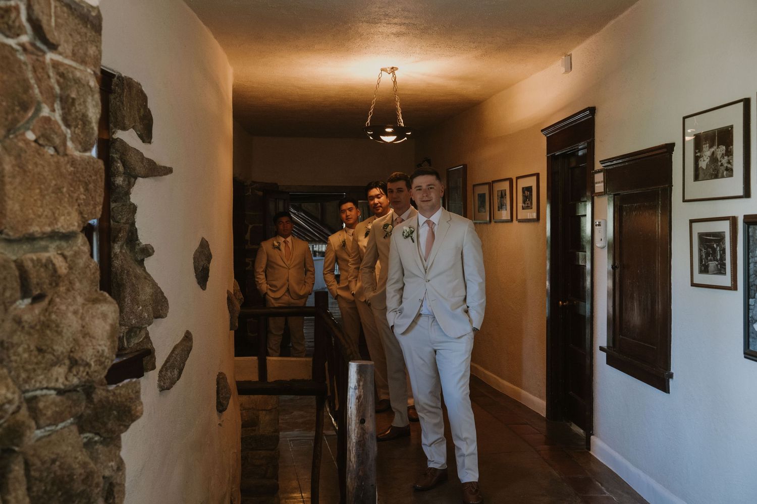 Groom and groomsmen in suits walking down a rustic hallway with stone walls and framed photos at a wedding venue.