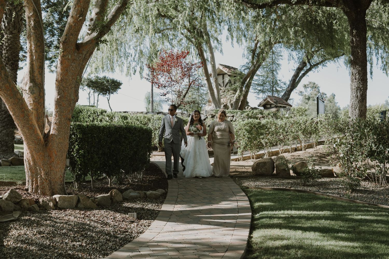 Bride in white gown walks garden path with two escorts toward outdoor wedding ceremony.