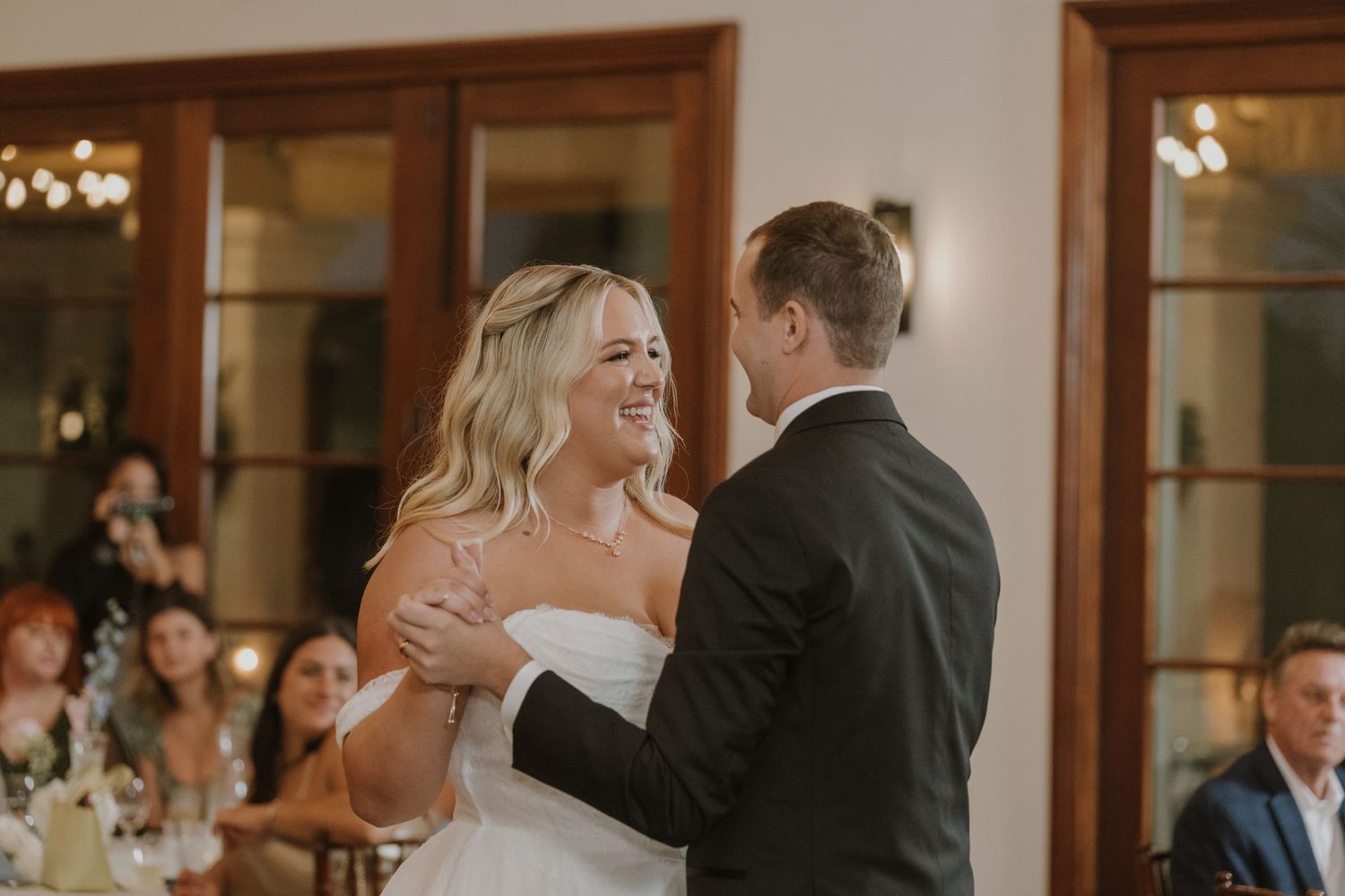 A smiling bride in a strapless white gown shares her first dance with the groom at their wedding reception.