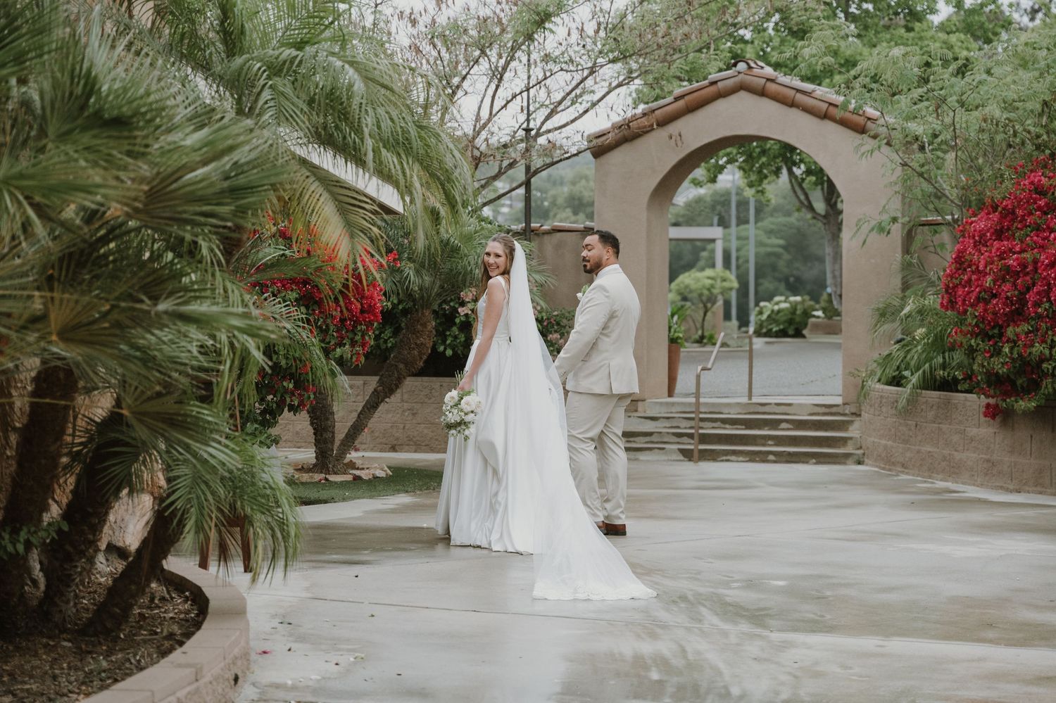 Bride and groom walk hand in hand through elegant outdoor venue with tropical palms and stone archway.