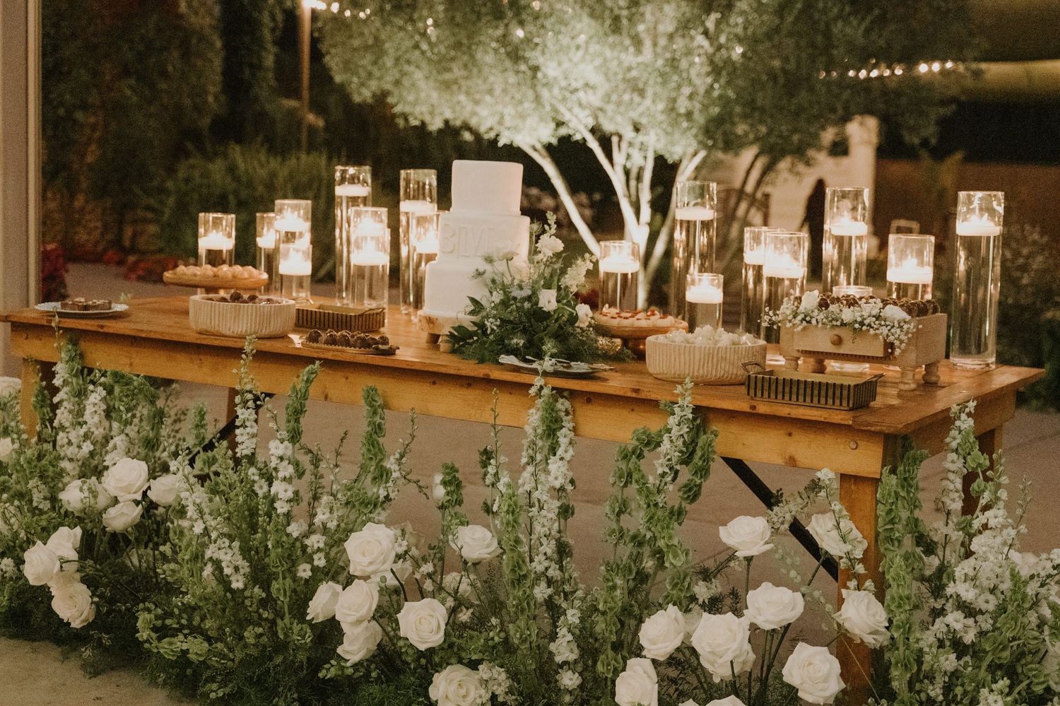 Romantic outdoor wedding dessert table adorned with white florals, candles, and string lights at night.