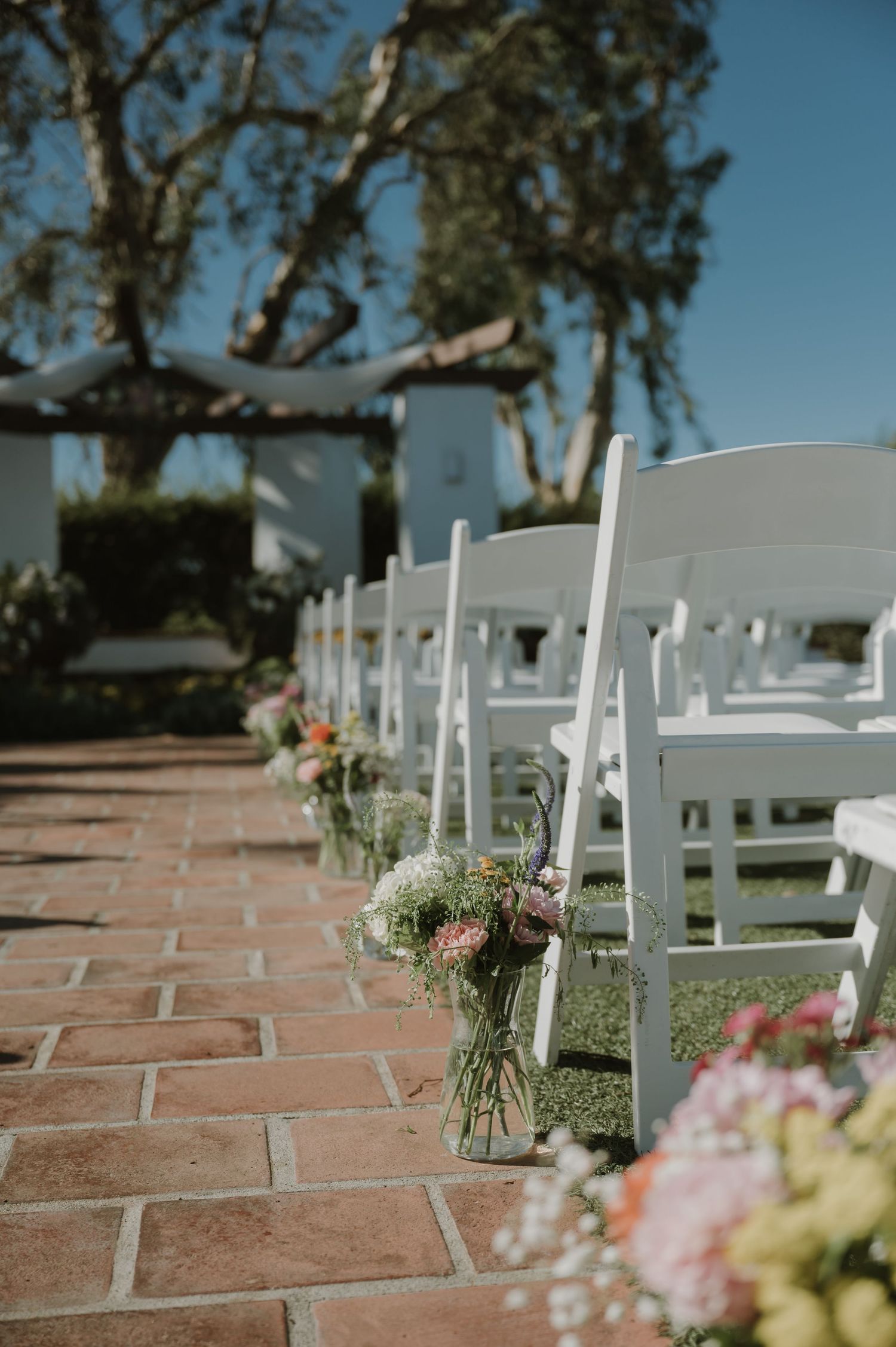 White folding chairs line a brick aisle adorned with mason jar wildflower arrangements at an outdoor wedding ceremony.