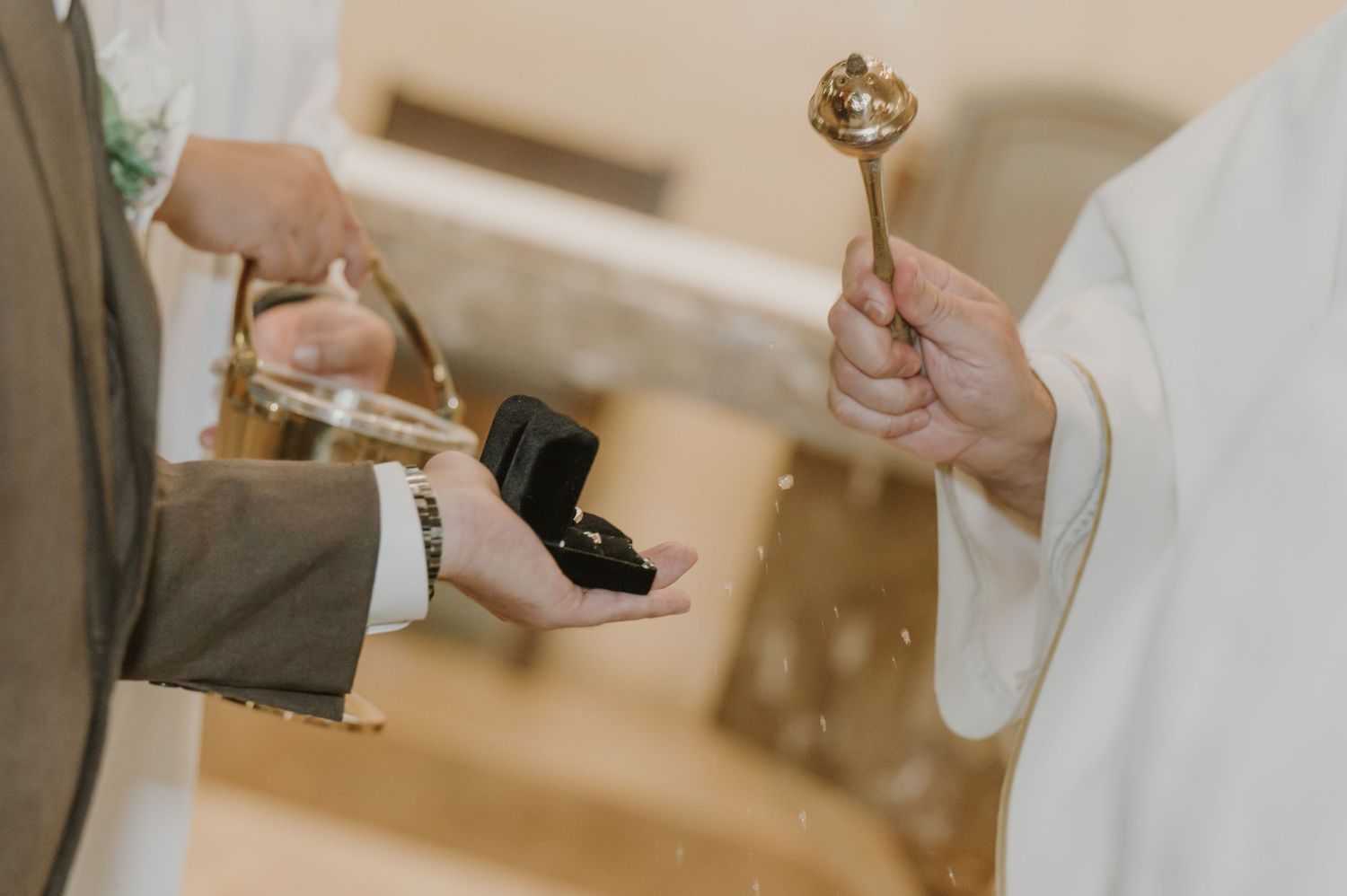 Priest blessing wedding rings with a golden aspergillum during a church ceremony.