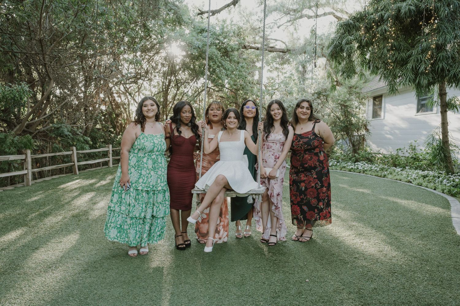 Bride in white dress being lifted by friends in colorful maxi dresses on garden pathway.