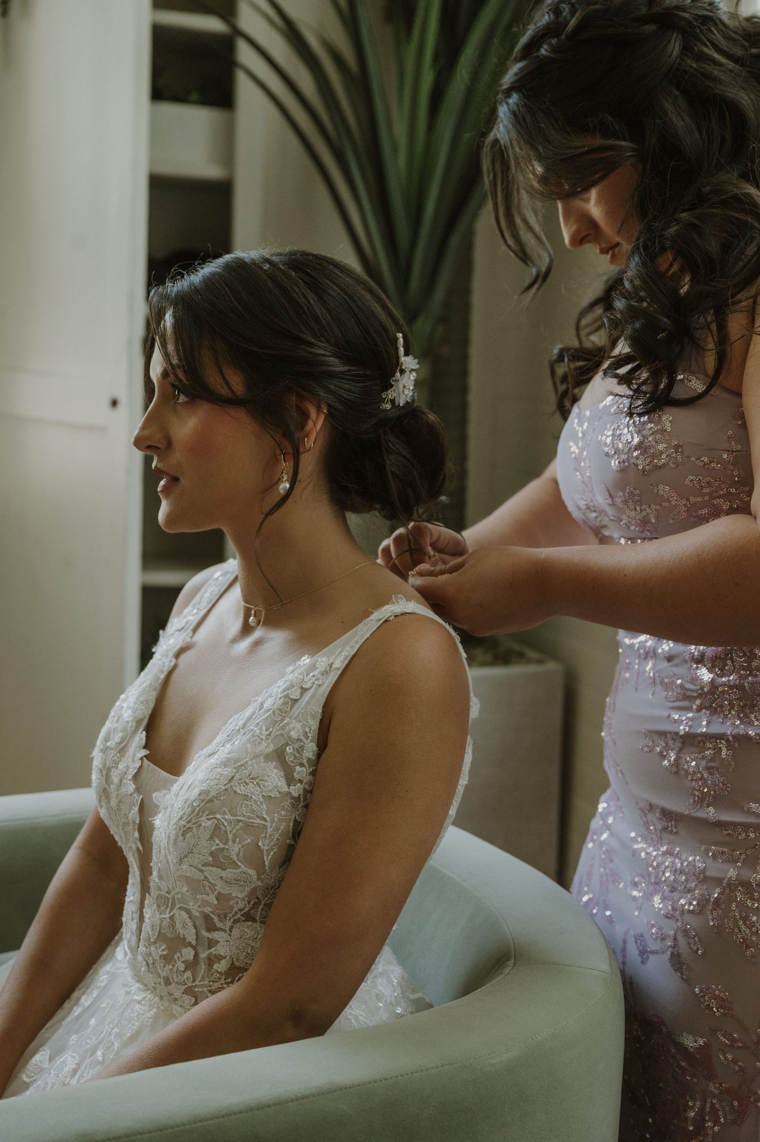 Bridesmaid in sequin dress fastening necklace on seated bride in lace gown before wedding ceremony.