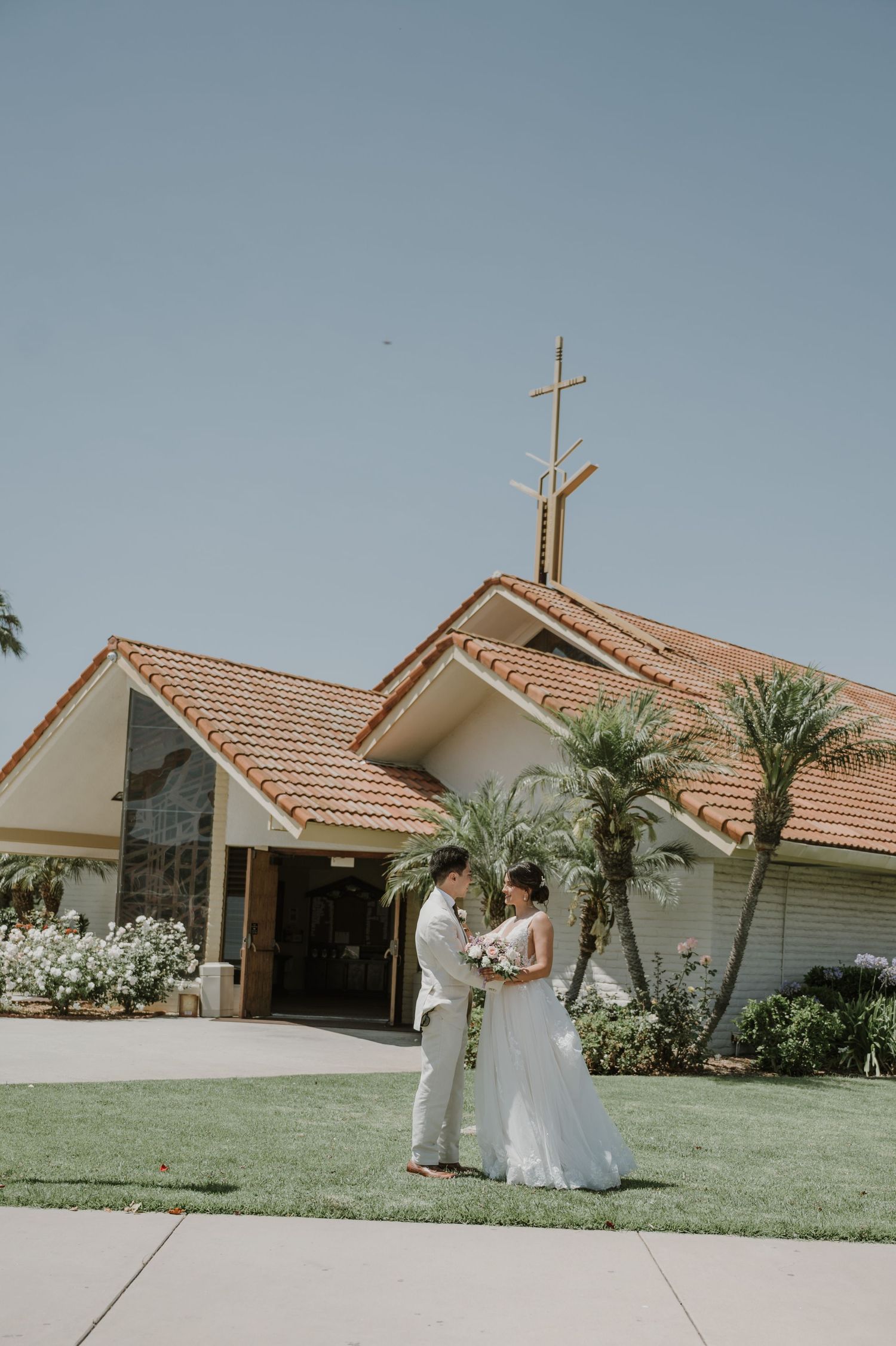 Bride and groom stand together on green lawn outside a church with terracotta roof and cross during outdoor wedding ceremony.