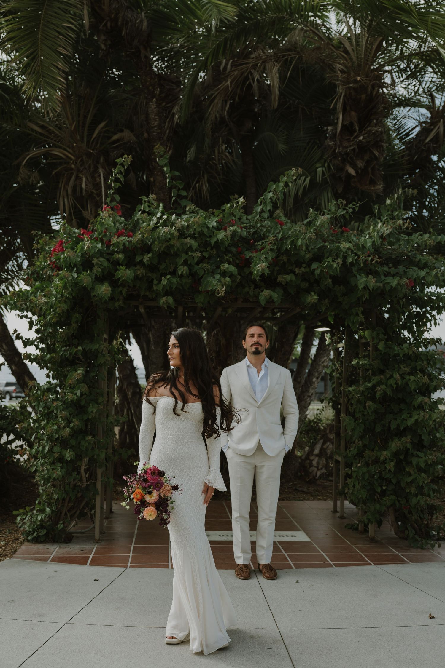 Wedding couple poses in white attire against lush tropical garden backdrop.