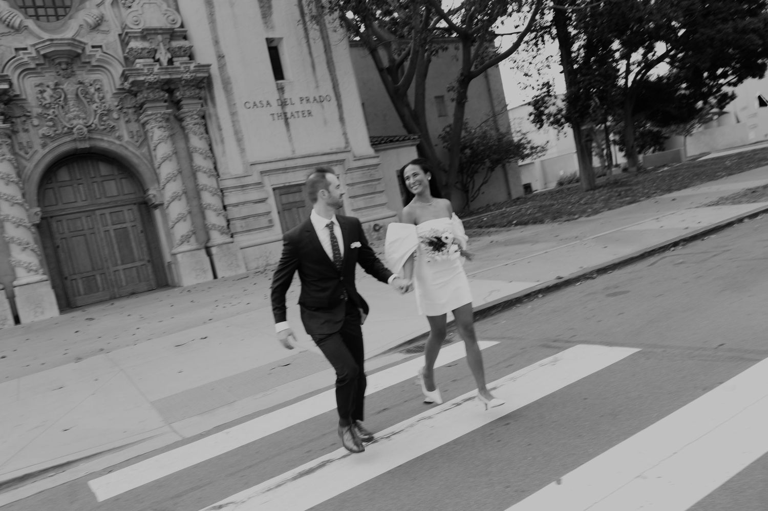 Wedding couple walks along a sidewalk in front of historic architecture.