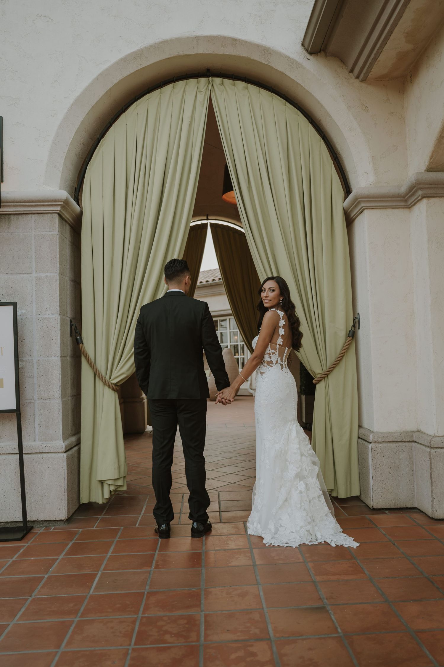 A wedding couple walking through an elegant arched hallway with sage green curtains.