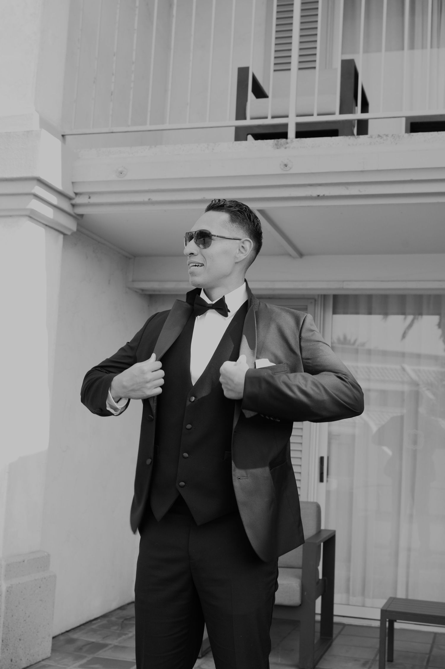 Black and white photo of groom in tuxedo and sunglasses posing confidently on balcony.