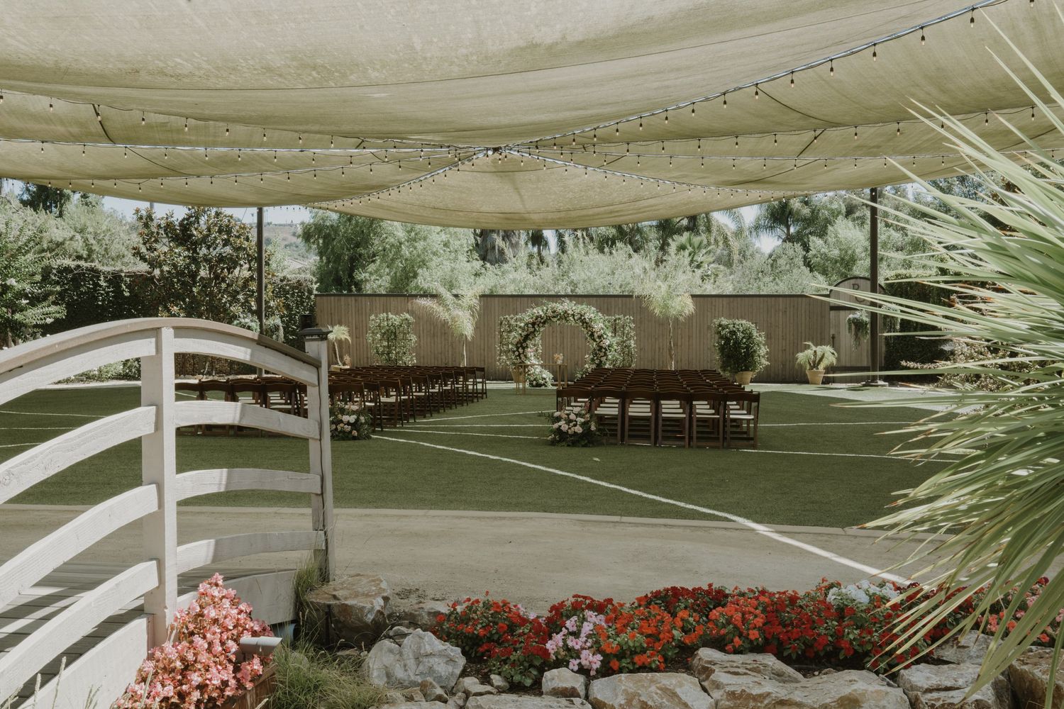 Outdoor wedding ceremony setup with floral arch, draped canopy, wooden chairs, and colorful garden flowers.
