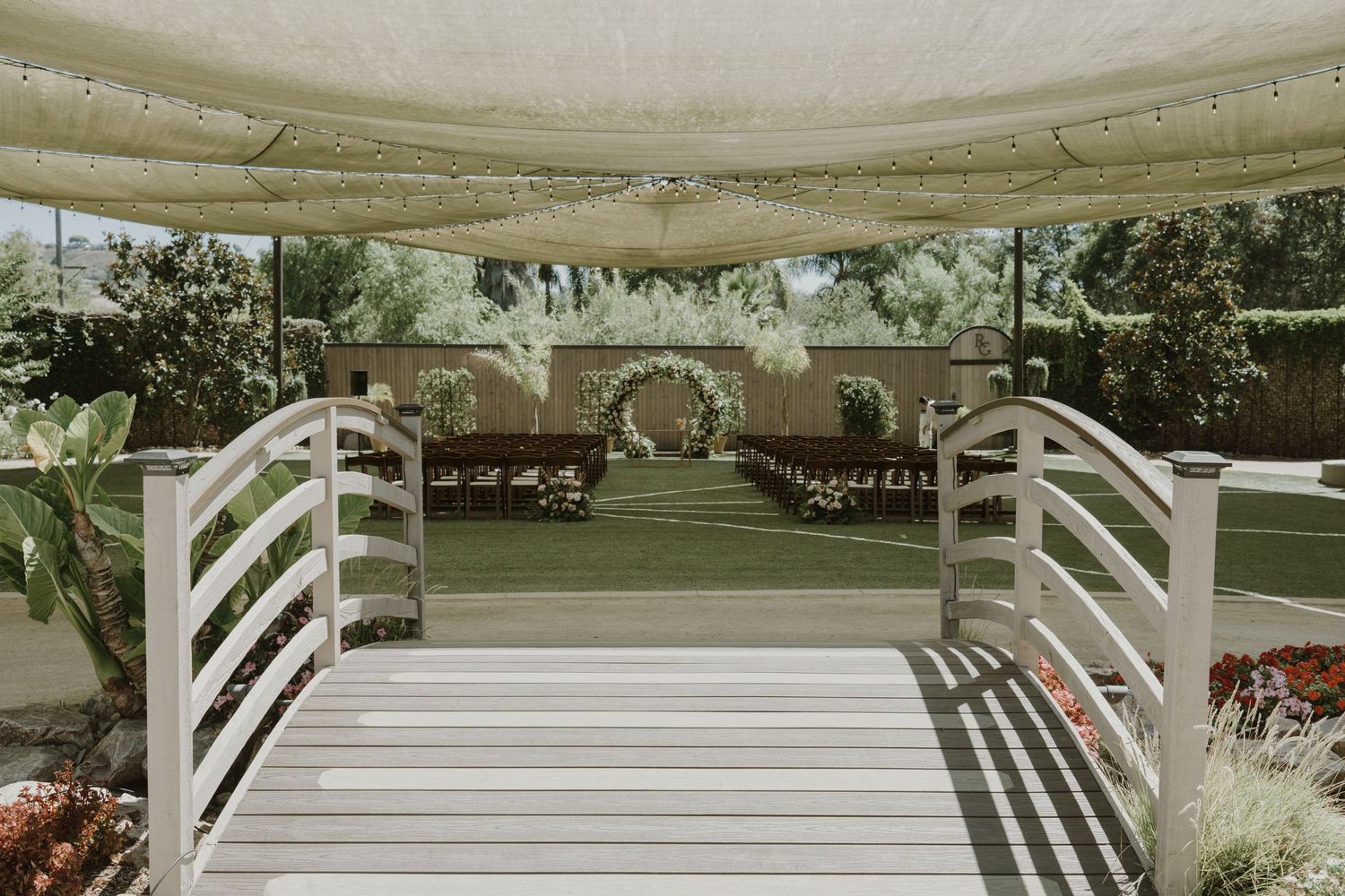 Elegant outdoor wedding ceremony setup with white wooden bridge, circular floral arch, and rows of chairs on green lawn.