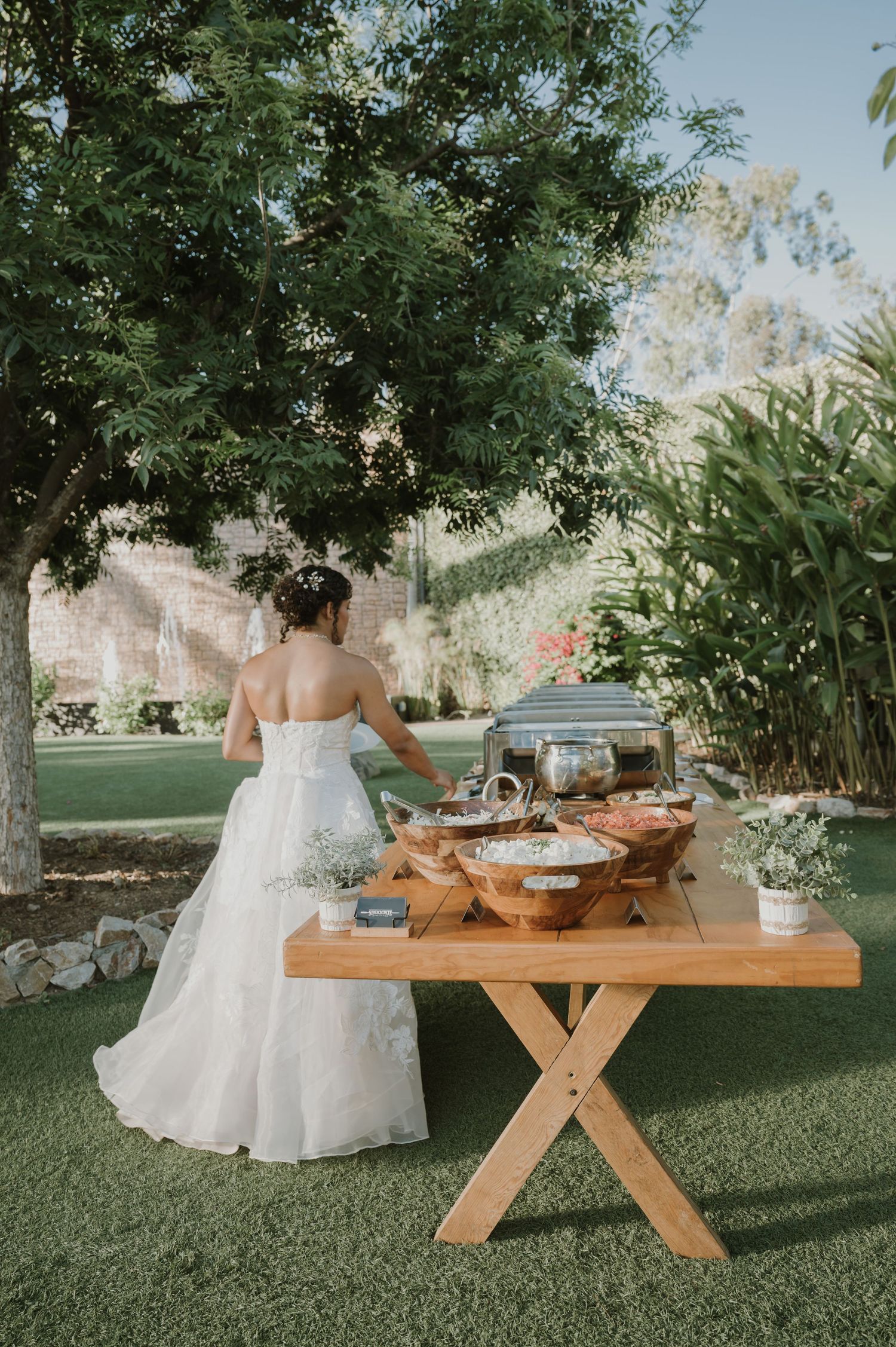 Bride in strapless lace gown serving food at outdoor wedding reception buffet table.