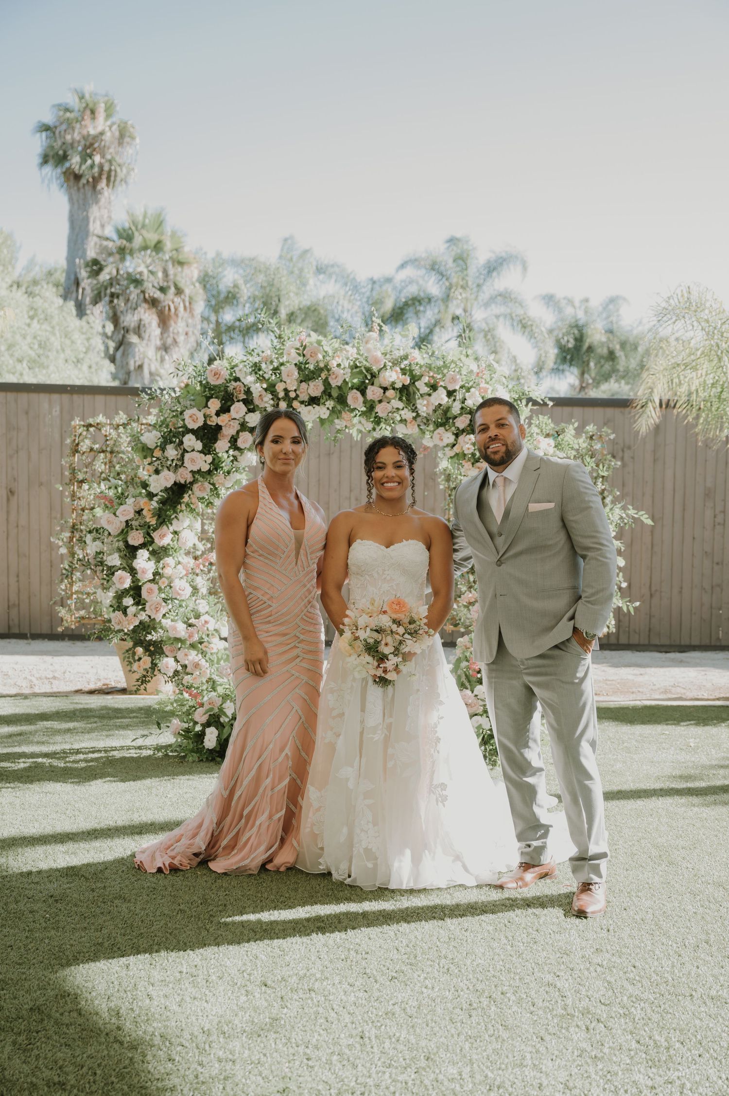 Bride in white gown poses with bridesmaid and groom before a floral arch at an outdoor wedding ceremony.