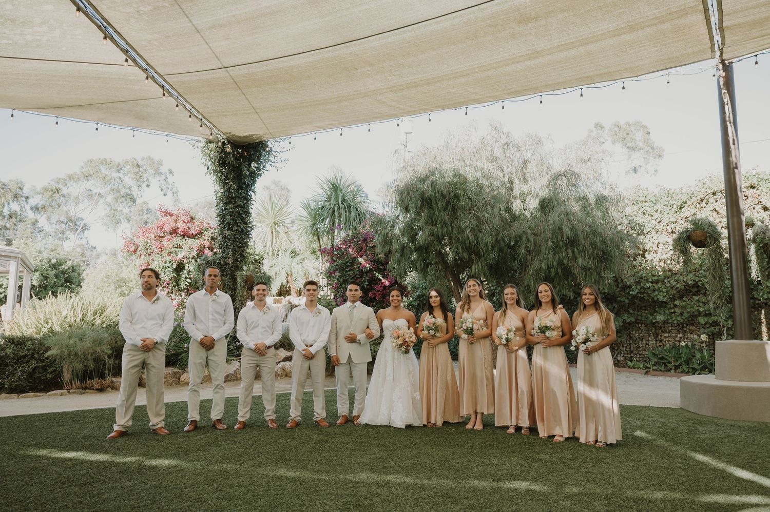 Wedding party in champagne and white attire poses together on lawn under draped canopy with string lights.