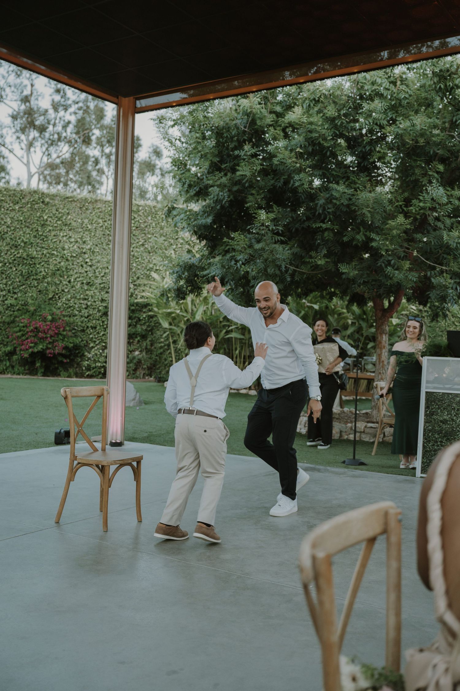 A man and young boy share a joyful dance together at an outdoor wedding reception under a covered patio.