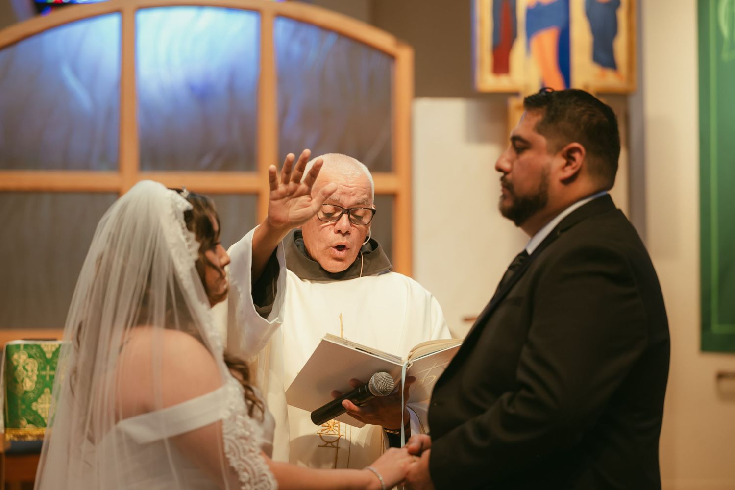 Elderly priest raises hand in blessing over bride and groom during wedding ceremony inside a colorful church.