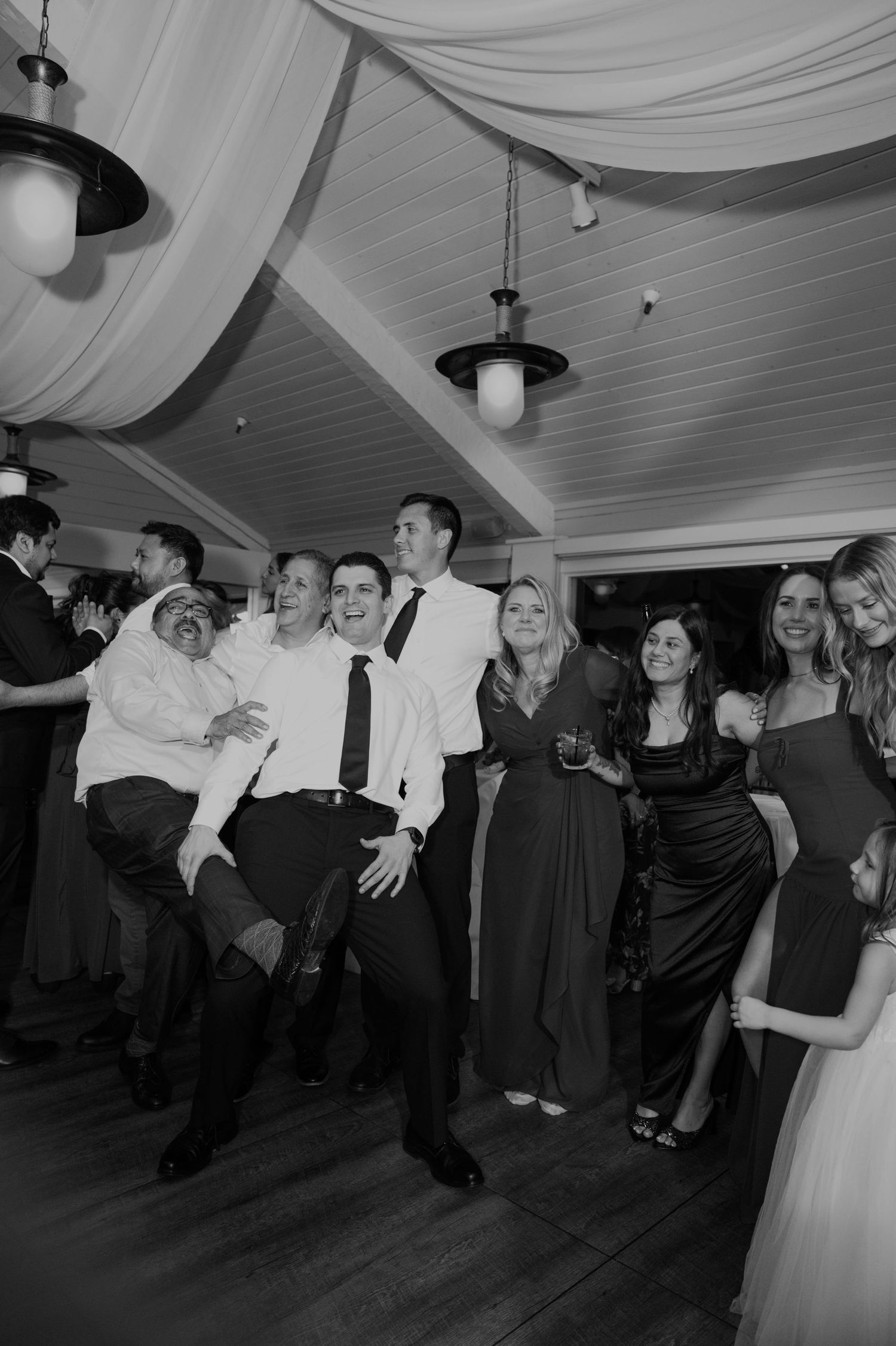Black and white photo of wedding guests laughing and dancing together at a lively reception.