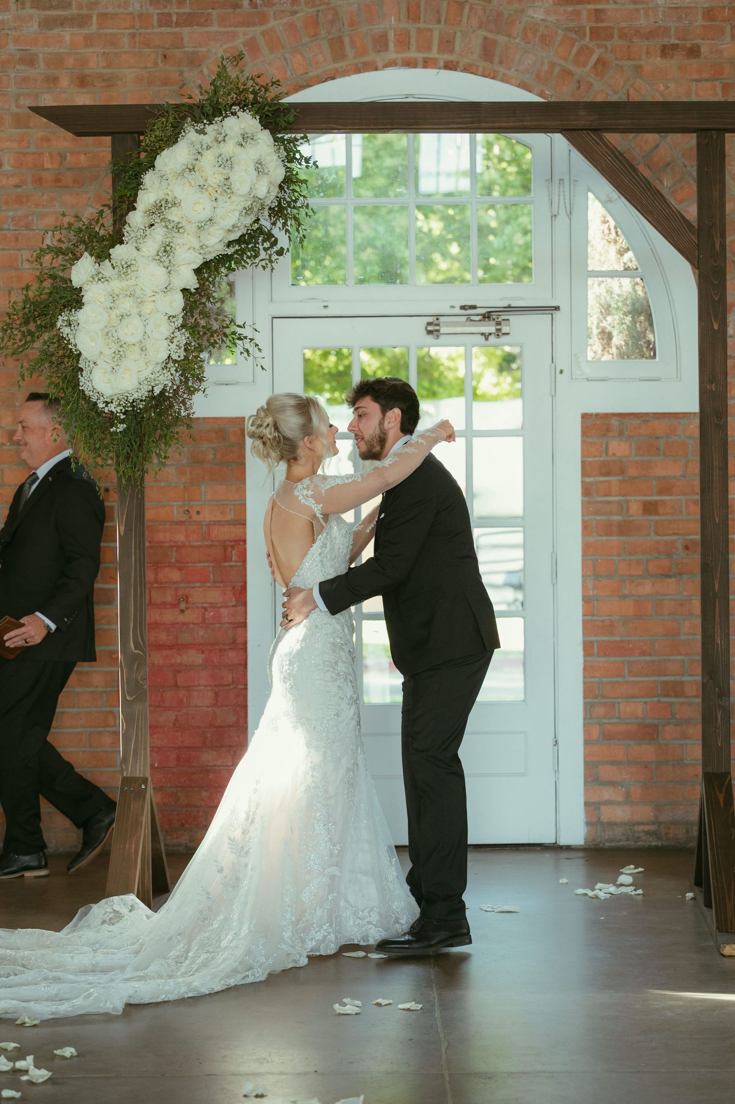 Bride and groom share first kiss under floral arch at indoor wedding ceremony in brick venue.