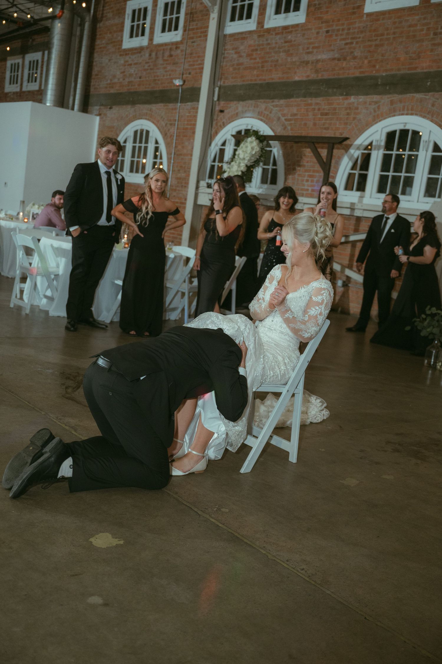 Groom removes bride's garter while she sits in chair surrounded by cheering wedding guests.