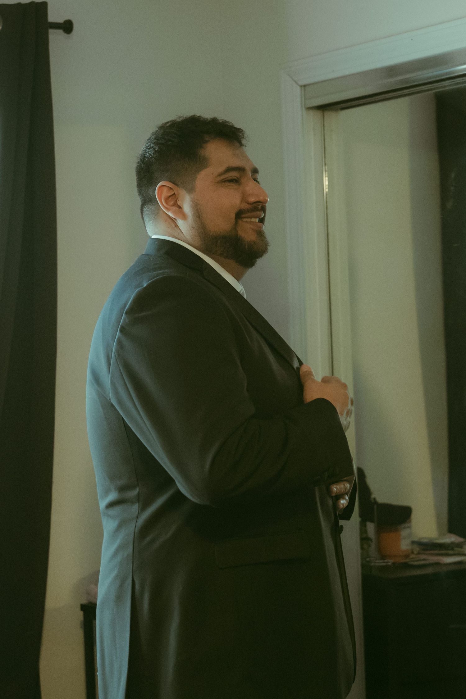 Smiling bearded man in a black suit adjusting his jacket indoors near a mirror.