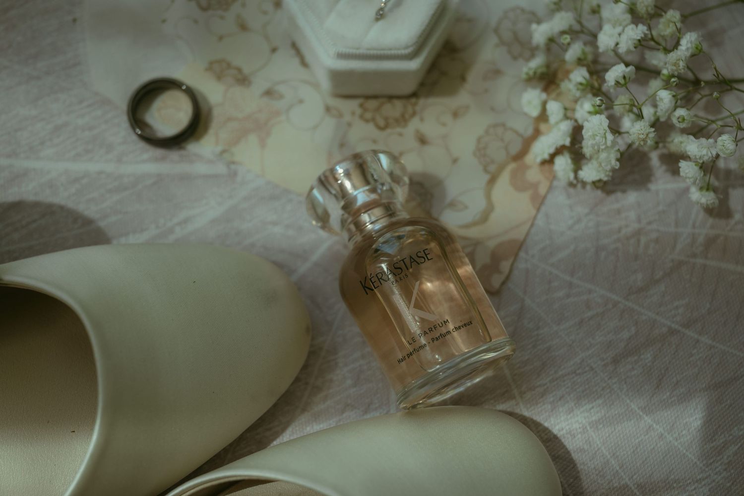 Bridal flat shoes, perfume bottle, ring, and white flowers arranged on a delicate floral fabric for a wedding.
