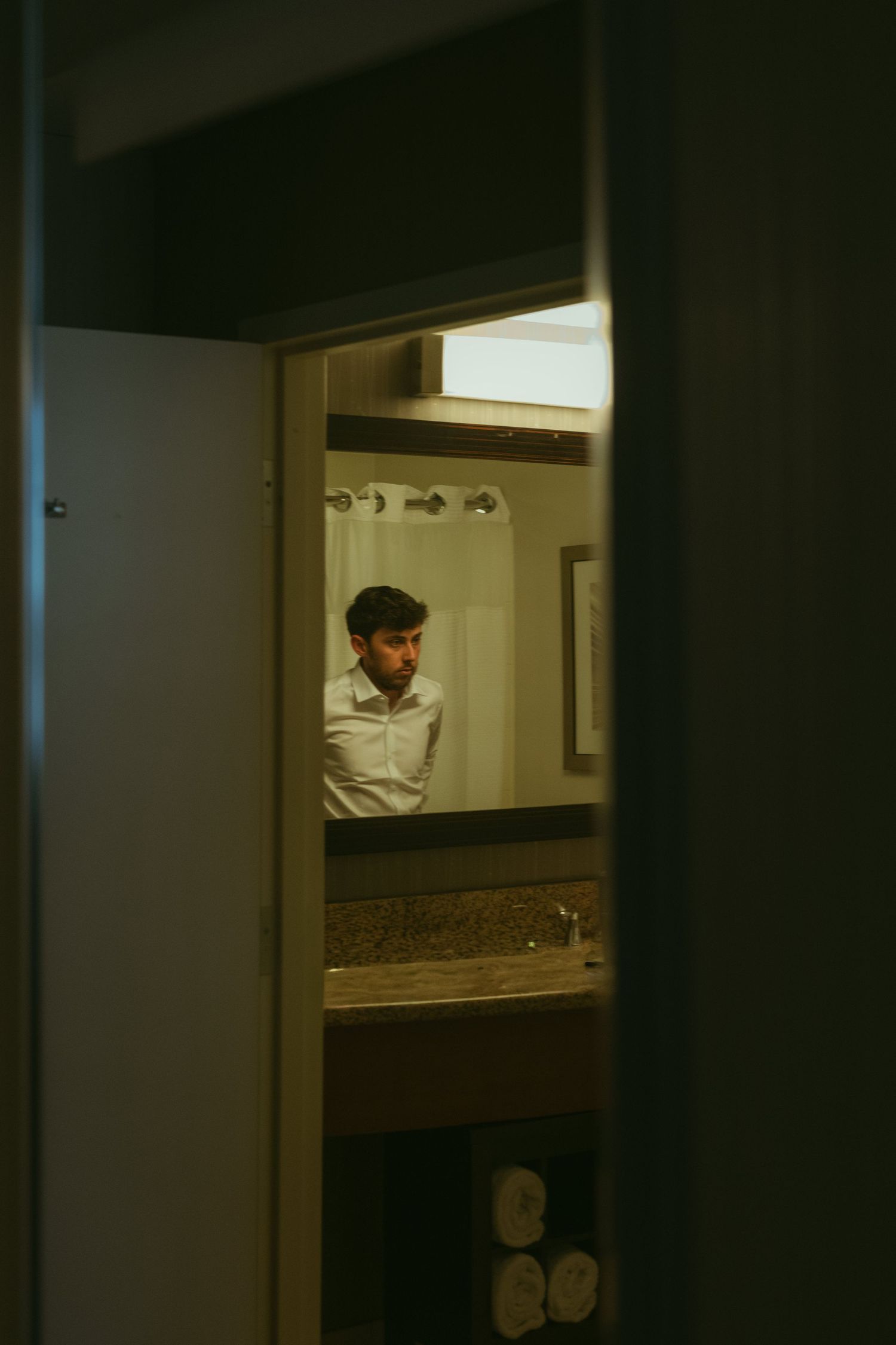 Groom in white dress shirt preparing in hotel bathroom mirror before wedding ceremony.