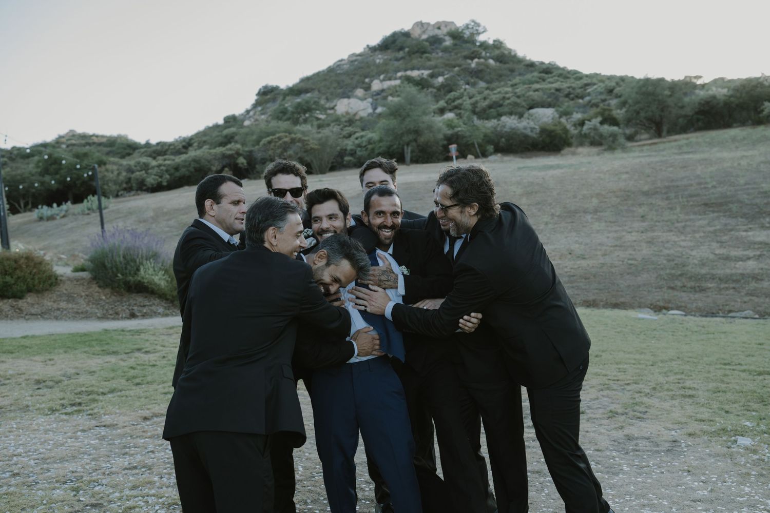 Groomsmen in dark suits laughing and hugging outdoors with green hills in background.