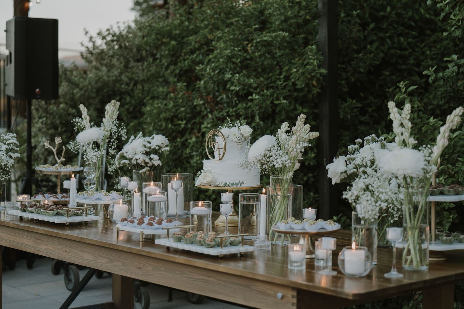 Elegant wedding dessert table with white floral arrangements, candles, and tiered cake on rustic wood table outdoors.