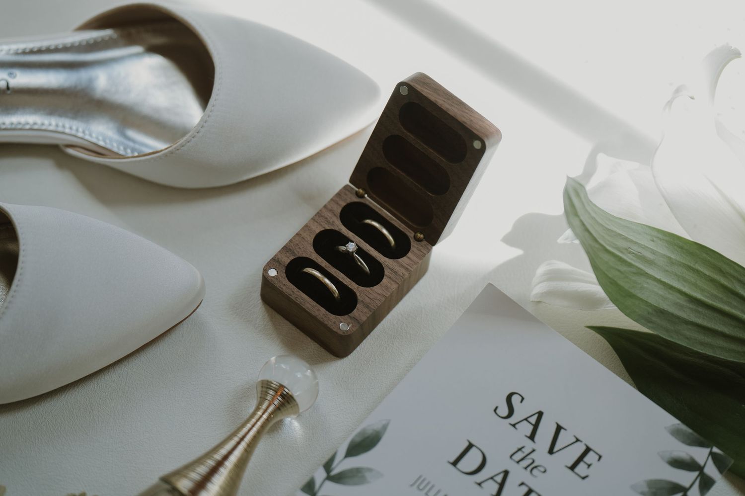 Wooden ring box with wedding bands beside white bridal heels and a Save the Date card on a white surface.