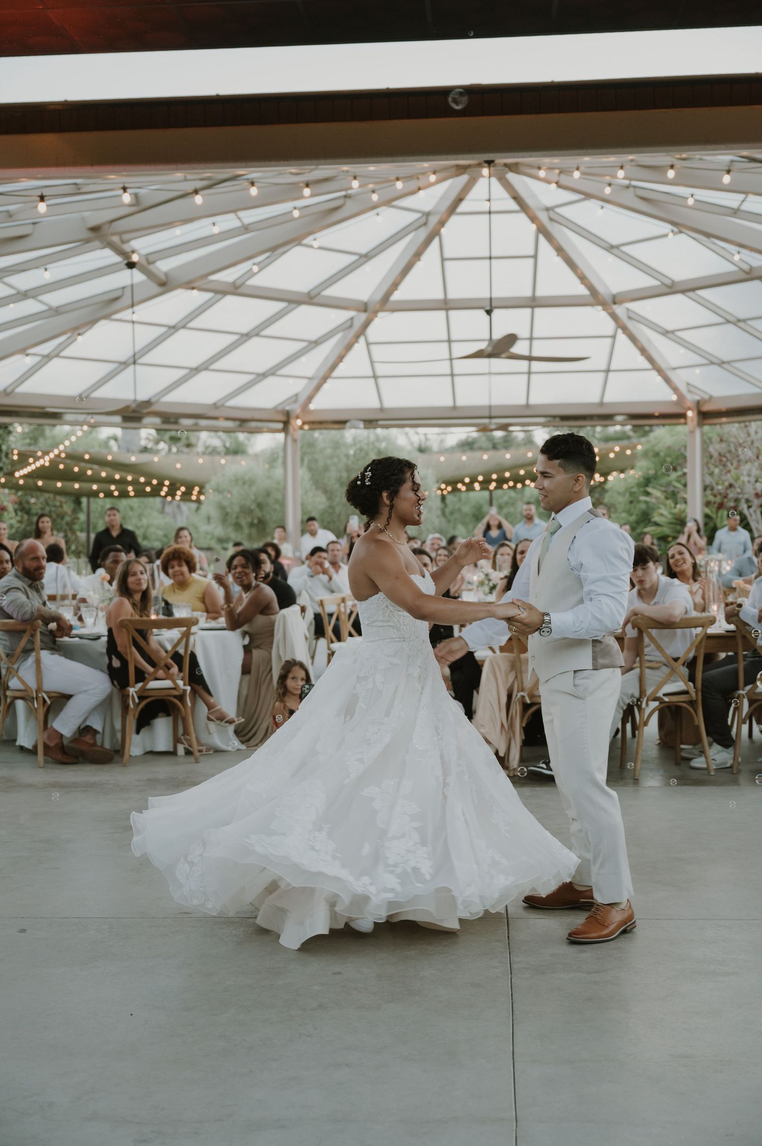 Bride and groom dance under string lights in elegant pavilion while wedding guests watch and celebrate.