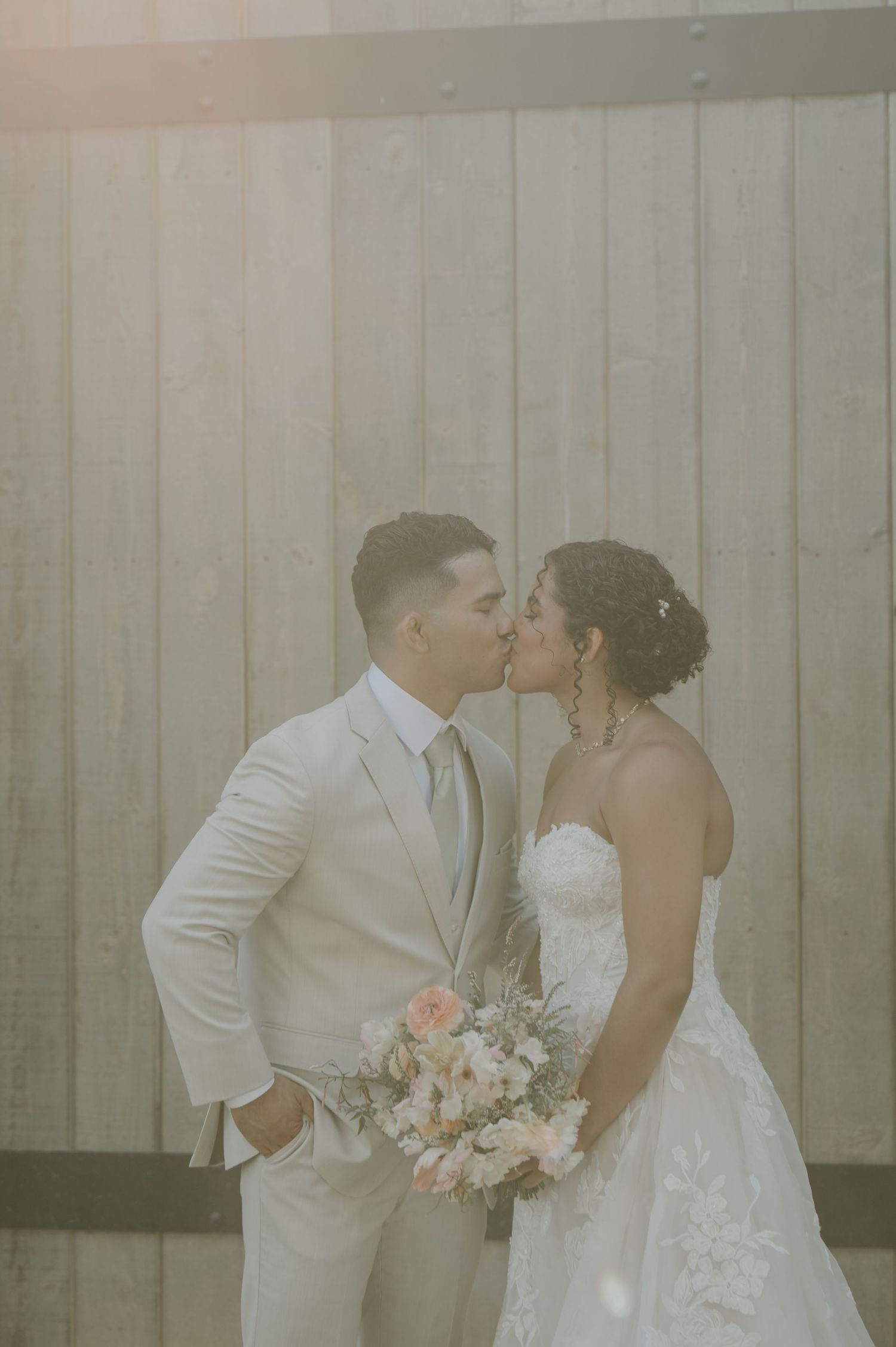 Bride and groom sharing a kiss in matching white wedding attire, bride holding a blush floral bouquet.