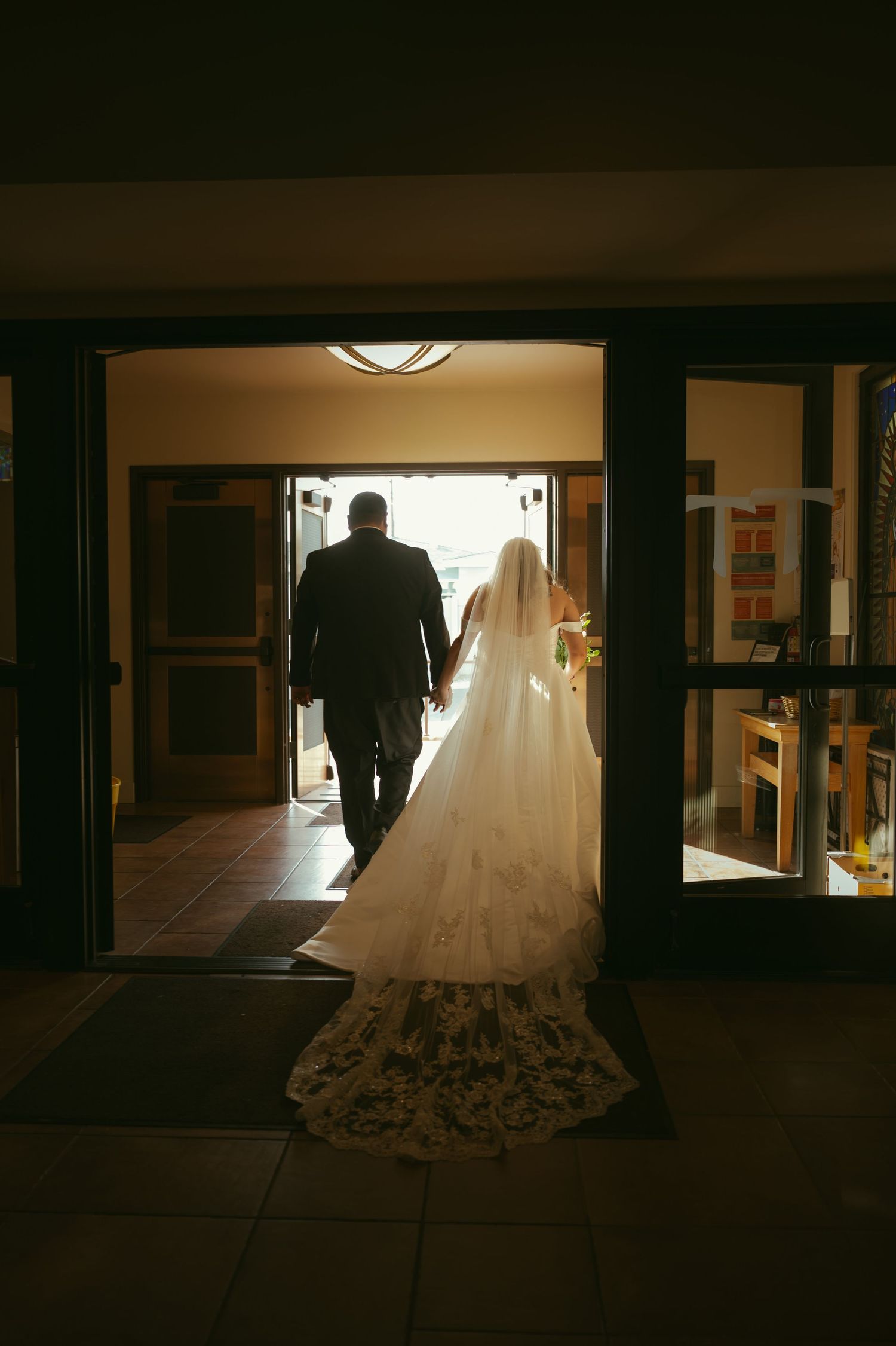 Silhouette of bride in lace gown and groom walking through church doors toward bright light.
