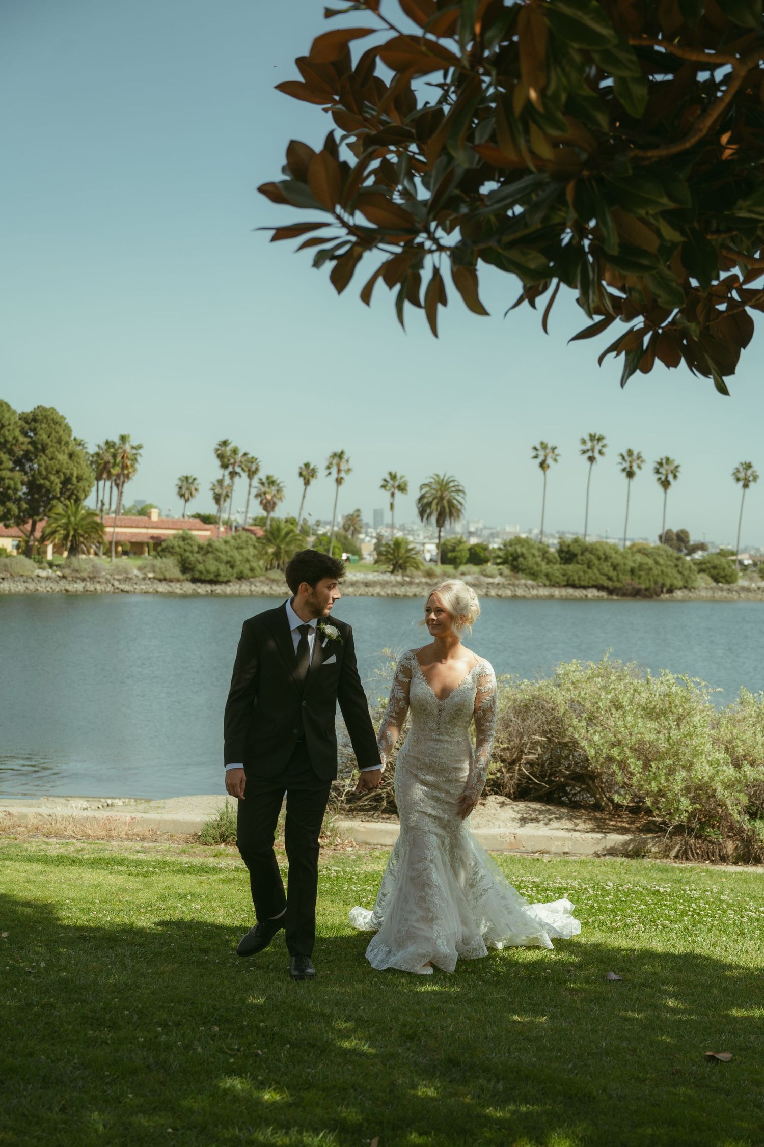 Bride and groom walking hand in hand on green lawn with palm trees and lake backdrop on wedding day.
