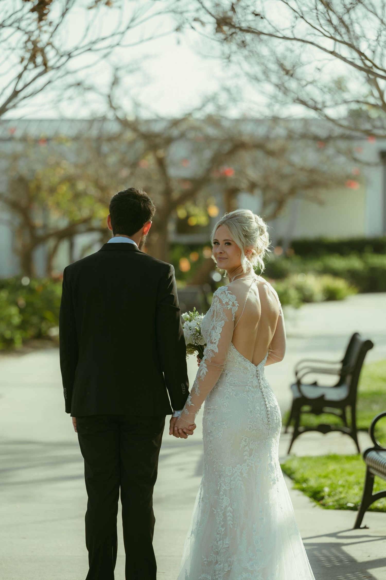 Bride in lace backless gown holds groom's hand, glancing back over shoulder in sunlit garden path.