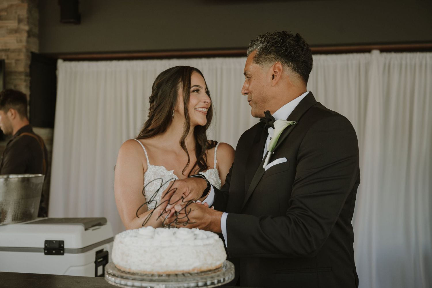 Bride and groom cutting white wedding cake together, smiling at each other in elegant venue.