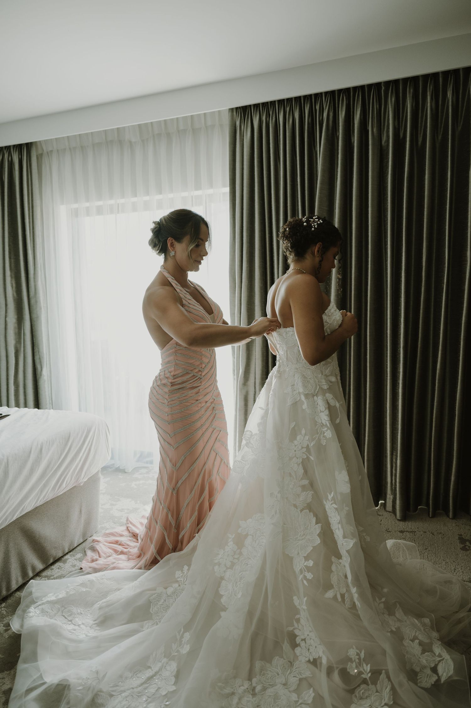Bridesmaid in pink gown helping bride with lace wedding dress in elegant hotel room.