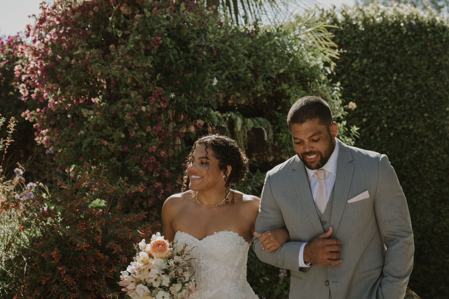 Happy bride and groom walking together outdoors, bride in strapless lace gown holding bouquet, groom in gray suit.