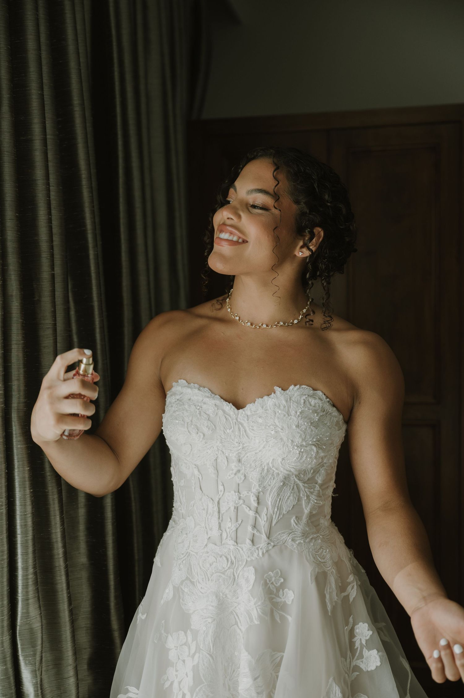 Smiling bride in a strapless lace wedding gown spraying perfume while getting ready.