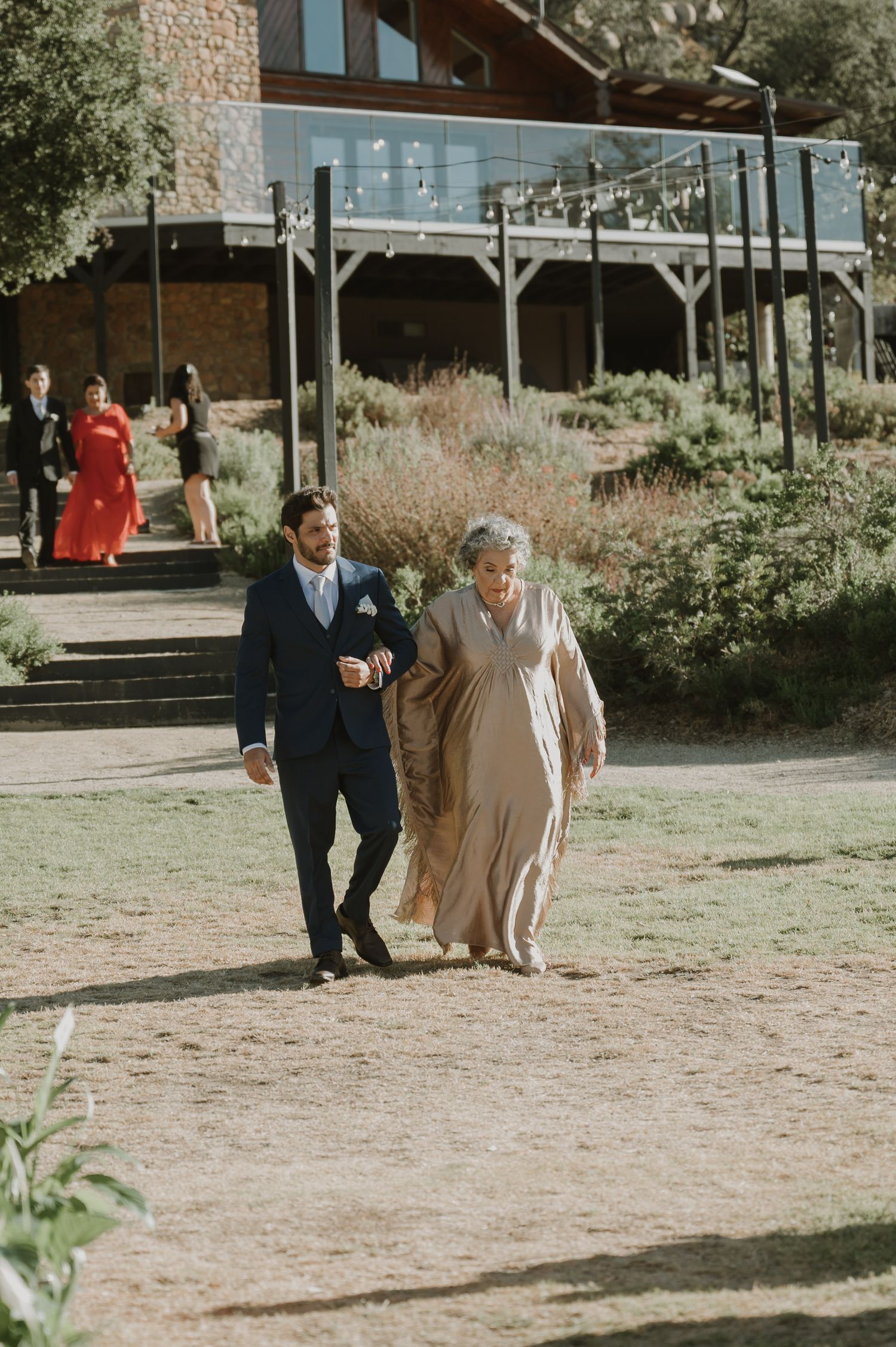 Groom in navy suit escorts elderly woman in beige gown along outdoor wedding venue path.