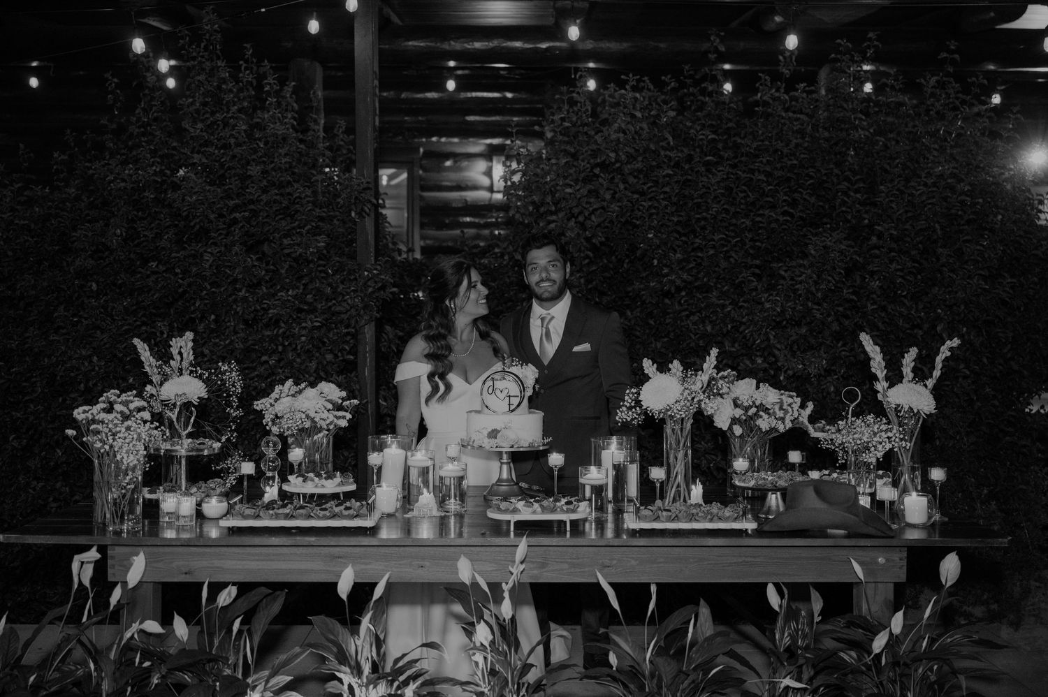 Bride and groom stand behind decorated sweetheart table with floral arrangements at outdoor wedding reception.