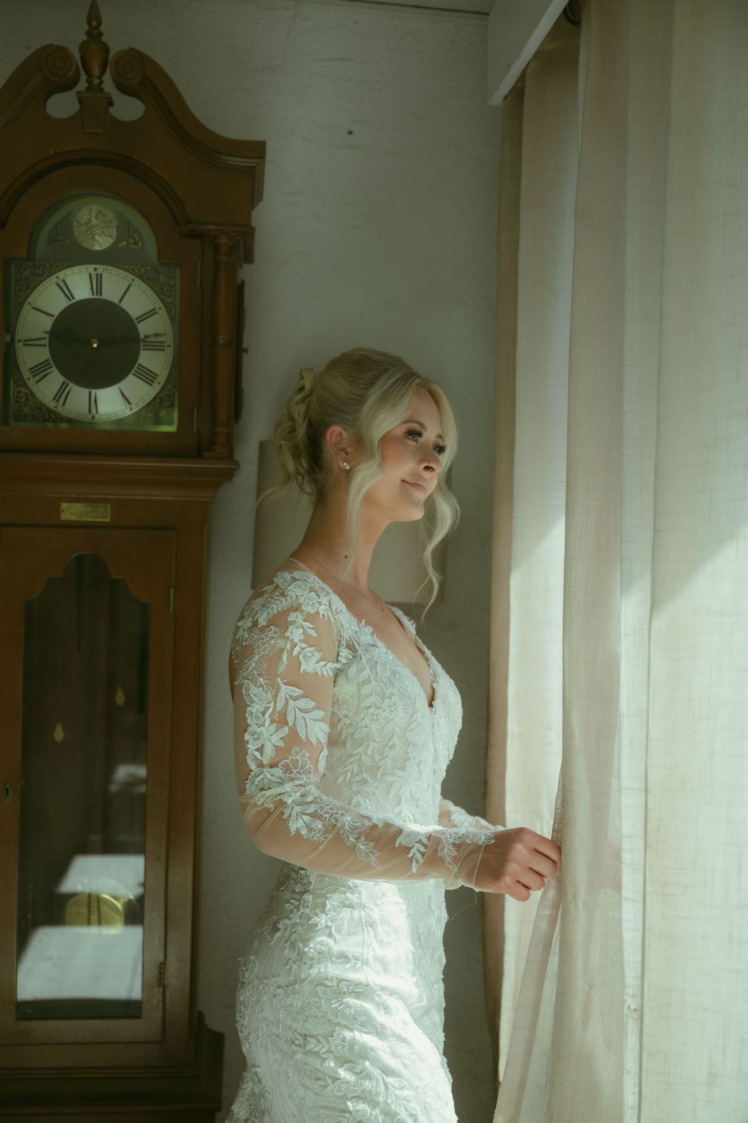 Beautiful bride in a lace long-sleeve wedding dress standing by a window near a grandfather clock.