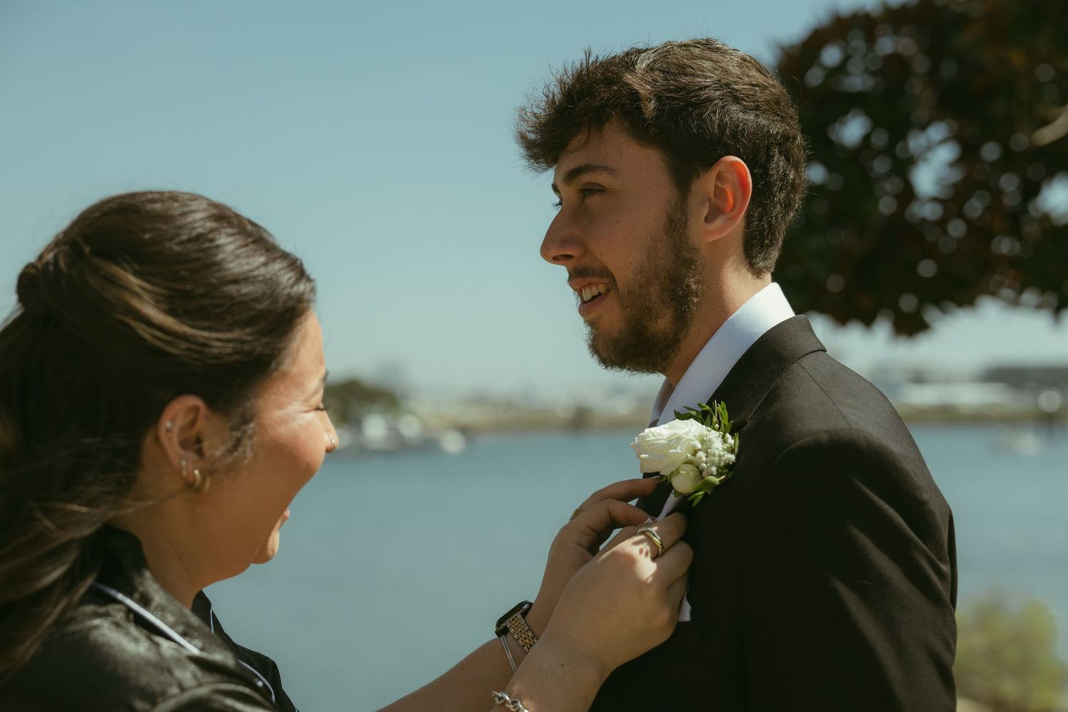 Woman pinning a white boutonniere on a smiling groom in a black suit outdoors near a waterfront.