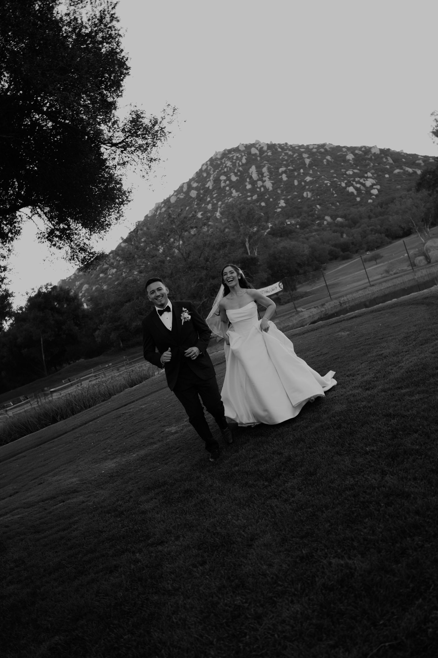 A joyful couple runs across a grassy field with mountains in the background at dusk.