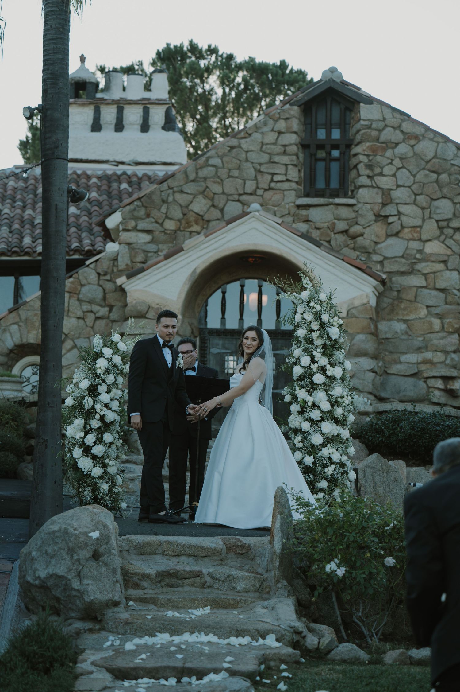 A wedding ceremony under a stone archway decorated with white flowers and greenery.